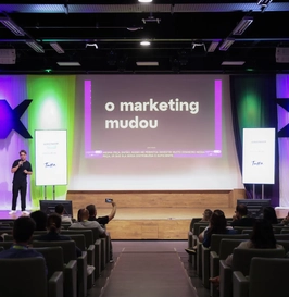 Foto de um auditório durante uma palestra. No centro do palco, o apresentador segura um microfone enquanto se dirige ao público. Ao fundo, um grande telão exibe sua apresentação com legendas acessíveis, garantindo a inclusão e a compreensão de todos os participantes.