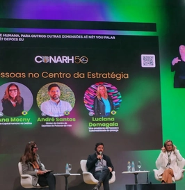 A imagem mostra um painel no evento CONARH 50 anos com o tema "Pessoas no Centro da Estratégia", destacando os palestrantes Ana Mocny, André Santos e Luciana Domagala, cujos nomes aparecem em um telão iluminado em verde. No palco, a moderadora e dois participantes estão sentados, enquanto uma intérprete de Libras traduz as falas e no telão estão geradas as legendas pelo Legenda.Live.