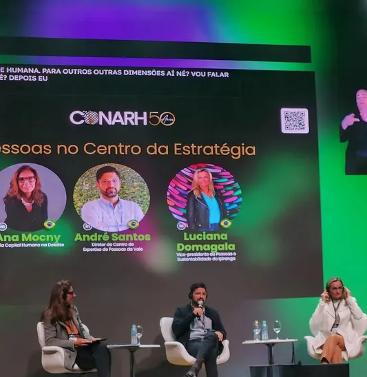 A imagem mostra um painel no evento CONARH 50 anos com o tema "Pessoas no Centro da Estratégia", destacando os palestrantes Ana Mocny, André Santos e Luciana Domagala, cujos nomes aparecem em um telão iluminado em verde. No palco, a moderadora e dois participantes estão sentados, enquanto uma intérprete de Libras traduz as falas e no telão estão geradas as legendas pelo Legenda.Live.