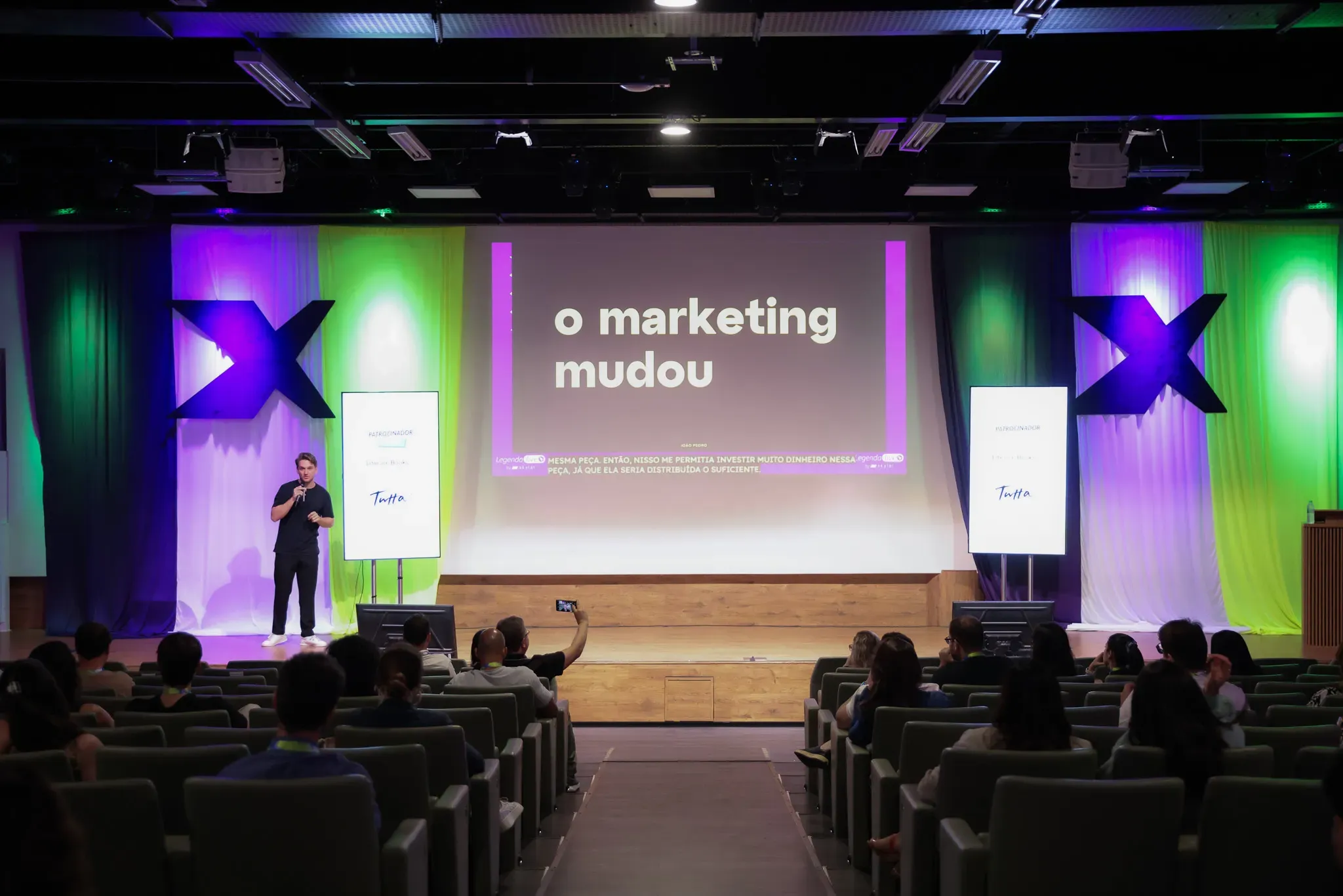Foto de um auditório durante uma palestra. No centro do palco, o apresentador segura um microfone enquanto se dirige ao público. Ao fundo, um grande telão exibe sua apresentação com legendas acessíveis, garantindo a inclusão e a compreensão de todos os participantes.