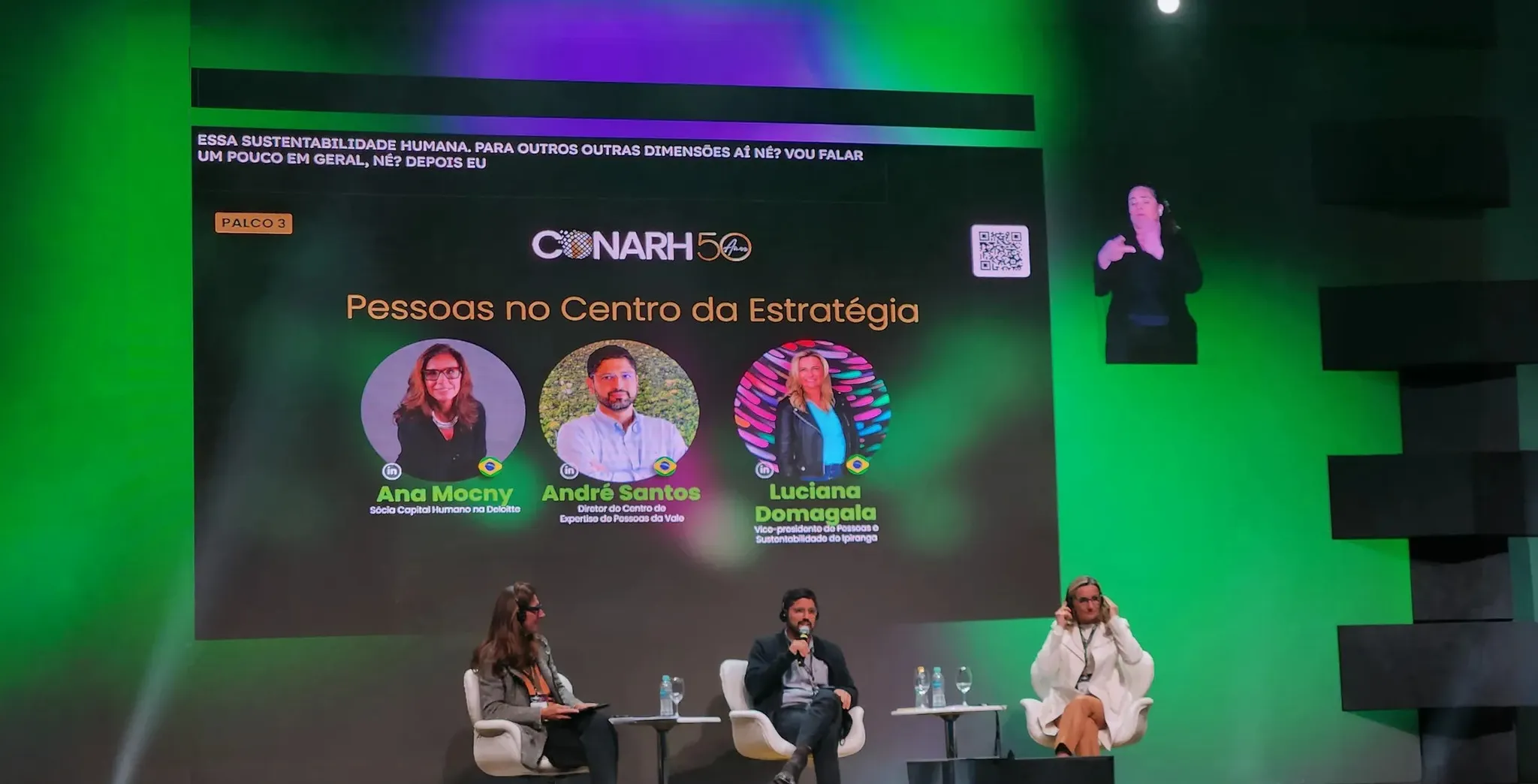 A imagem mostra um painel no evento CONARH 50 anos com o tema "Pessoas no Centro da Estratégia", destacando os palestrantes Ana Mocny, André Santos e Luciana Domagala, cujos nomes aparecem em um telão iluminado em verde. No palco, a moderadora e dois participantes estão sentados, enquanto uma intérprete de Libras traduz as falas e no telão estão geradas as legendas pelo Legenda.Live.